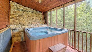 Eine Holz-Blockhütte mit einer Steinwand, einem Whirlpool und einem Balkon mit Blick auf Bäume.