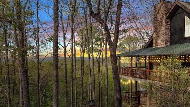 Uma cabana rústica com deck de madeira e luzes de cordão, cercada por árvores altas e um céu limpo.