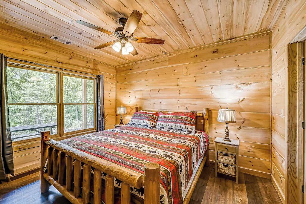 A wooden cabin bedroom with a bed, two bedside lamps, a ceiling fan, and a window.