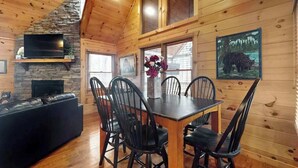 A wooden cabin interior with a dining table, chairs, a fireplace, and a television mounted on a stone wall. - Blue Ridge Bear Bungalow (Blue Ridge)