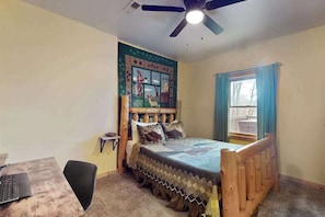 Ein Schlafzimmer mit einem Bett, einem Schreibtisch mit Laptop, einem Deckenventilator und einem Fenster mit Vorhängen.
