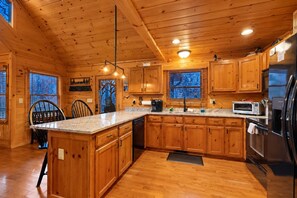 A wooden cabin interior with a kitchen featuring wooden cabinets, a granite countertop, and modern appliances. - Blue Ridge Bear Bungalow (Blue Ridge)