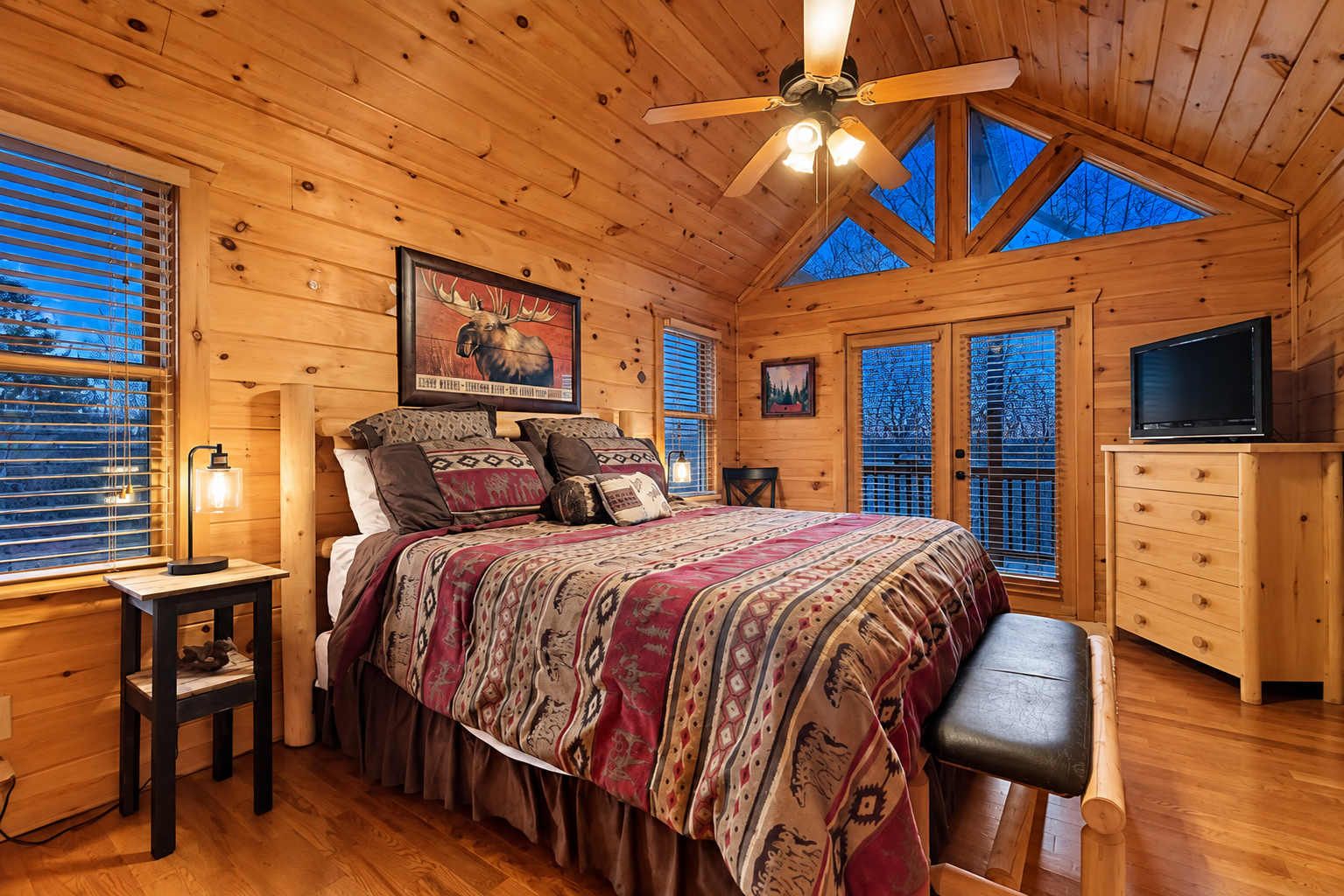A wooden cabin bedroom with a bed, a desk, a chair, a television, and a ceiling fan.