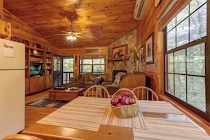 A cabin interior with a dining area, wooden walls, a fireplace, and large windows. - River Ranch Ellijay Stay (Ellijay)