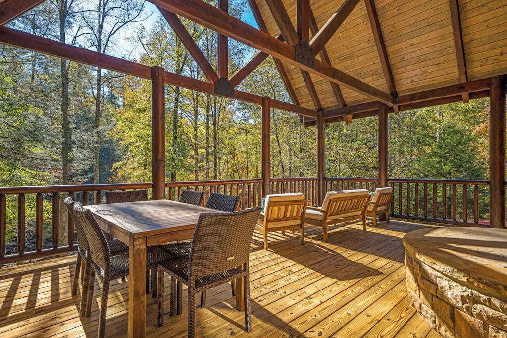 A covered deck with a table and chairs, surrounded by a wooden railing, overlooking a forest.