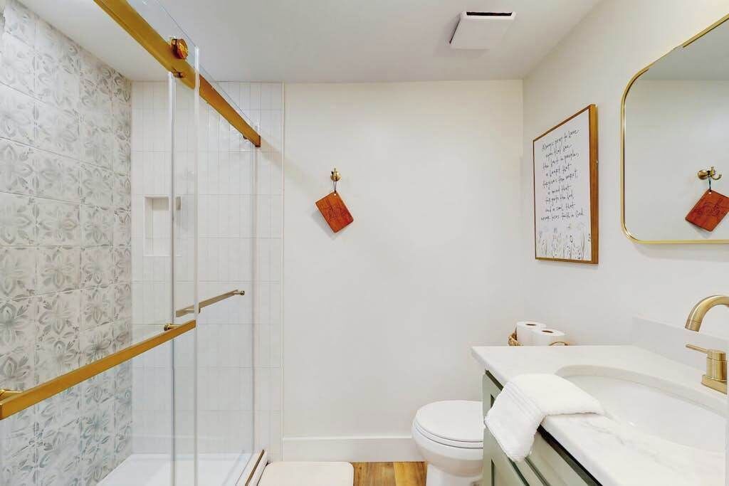 Ein Badezimmer mit einer Duschkabine aus Glas, einer weißen Toilette, einem Waschbecken mit goldener Armatur und einem Spiegel mit goldener Rahmung.