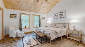 Ein Schlafzimmer mit Holzboden, einem Bett, einem Nachttisch, einem Sessel und zwei Fenstern.