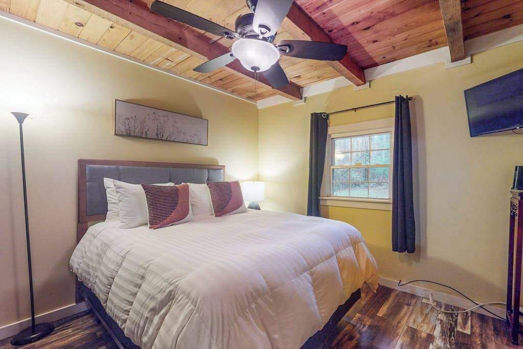 Een slaapkamer met een bed, plafondventilator, raam met gordijnen en een schilderij aan de muur.
