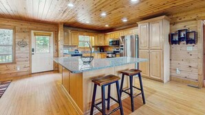 Interior de una cabaña de madera con cocina que incluye armarios de madera, una isla central con taburetes y electrodomésticos integrados.