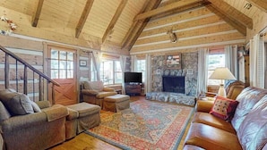 A log cabin interior with a stone fireplace, wooden ceiling beams, a television, and a cozy seating area. - Treehouse Retreat Stay (Sky Valley)