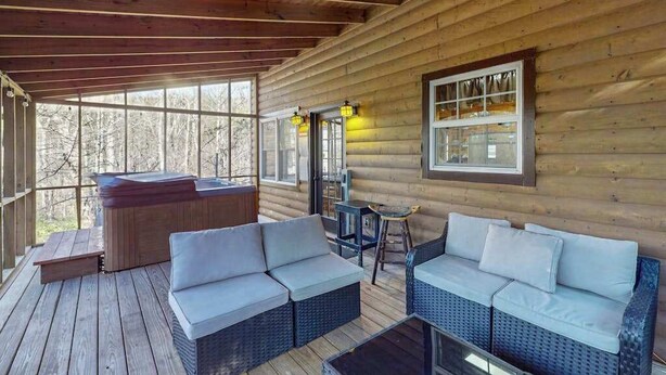 A wooden cabin with a covered porch, a hot tub, wicker furniture, and a window overlooking trees. - Bear's Den Sautee Cabin (Sautee Nacoochee)