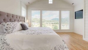 Een slaapkamer met een groot bed, een raam met zonwering, een plafondventilator en uitzicht op het omliggende landschap.