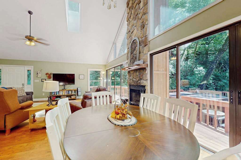 Amplia sala de estar con una gran mesa de madera, asientos cómodos, chimenea y ventanas grandes que ofrecen vistas al exterior.