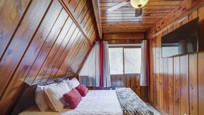 A bedroom with a sloped ceiling, a bed with pillows and a blanket, a television on the wall, and a window with curtains. - Paradise Retreat Mountainview (Jasper)
