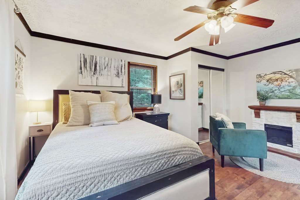 A bedroom with a bed, a ceiling fan, a fireplace, and artwork on the walls.