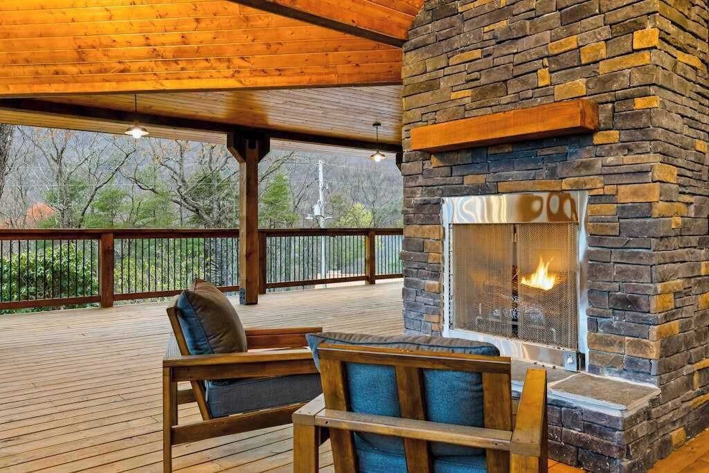 An outdoor deck with a stone fireplace, wooden furniture, and a view of trees.