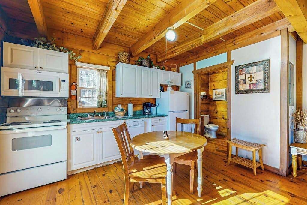 A wooden cabin interior with a kitchen area featuring white cabinets, a stove, a microwave, and a dining table with chairs.