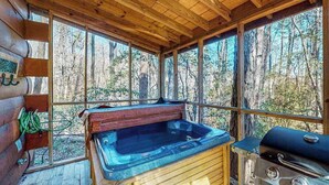 A cabin with a hot tub, a grill, and large windows overlooking a forest.