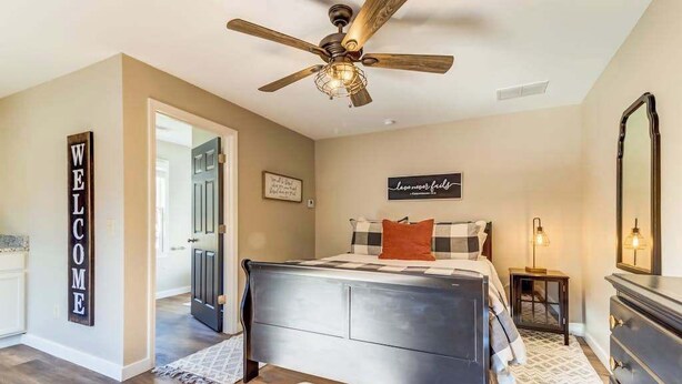 A bedroom with a bed, a ceiling fan, a dresser, a nightstand, and a wall sign.