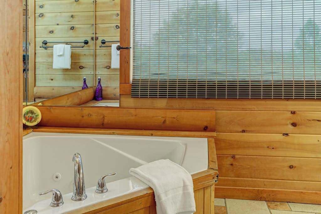 A wooden cabin with a bathtub, a window with blinds, and a mirror reflecting the interior.