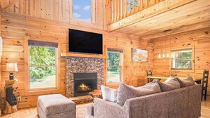 A log cabin interior with a stone fireplace, a flat-screen TV, a gray sofa, and a dining area. - River Fishing Cabin - Dog Friendly (Lakemont)