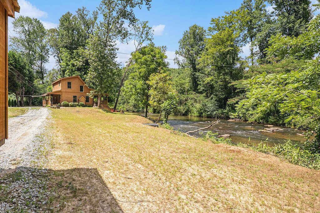 Een rustiek houten huis met een grindpad ernaartoe, omgeven door weelderige begroeiing en een rivier.