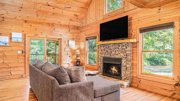 A log cabin interior with a stone fireplace, a large TV mounted above it, and a cozy seating area. - River Fishing Cabin - Dog Friendly (Lakemont)