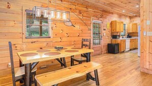 A wooden cabin interior with a dining table, chairs, a bench, a kitchen area, and a hanging light fixture. - River Fishing Cabin - Dog Friendly (Lakemont)
