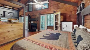 A log cabin interior with a fireplace, a bed with a buffalo print, a ceiling fan, and a kitchen area.