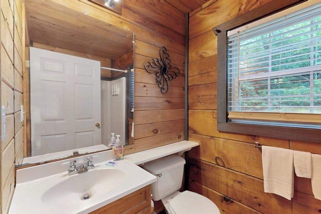 A wooden cabin bathroom with a white sink, toilet, and window with blinds.