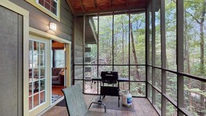 A covered porch with a bench, a grill, and a large window overlooking a forest.