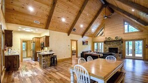 A spacious kitchen with wooden cabinets, a dining area with a large wooden table and white chairs, and a stone fireplace. - Toccoa River Run Cabin (Mineral Bluff)
