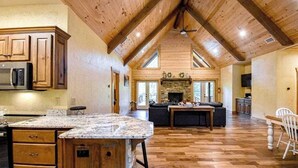 A spacious kitchen with wooden cabinets, a granite countertop island, and a stone fireplace. - Toccoa River Run Cabin (Mineral Bluff)