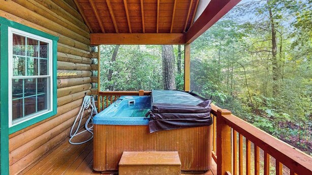 Una cabaña de troncos con una terraza de madera, un jacuzzi y una ventana con vistas a un bosque.