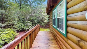 Una cabaña de troncos con una terraza de madera que lleva a una ventana, rodeada de un bosque frondoso.