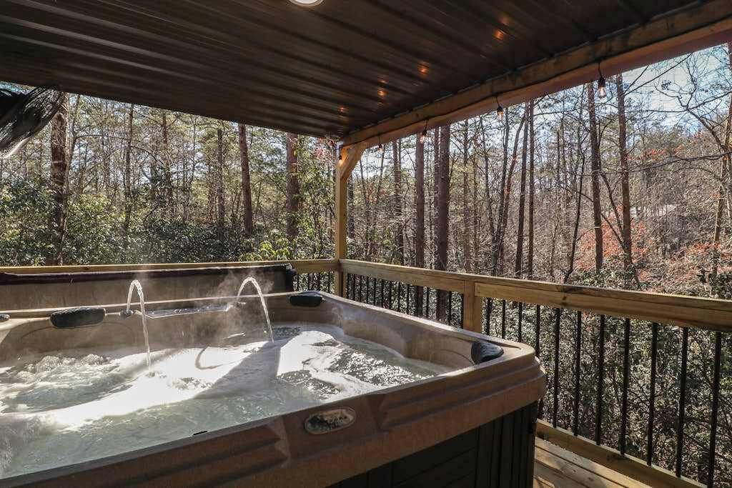 A hot tub with a wooden deck and railing, surrounded by a forested area.