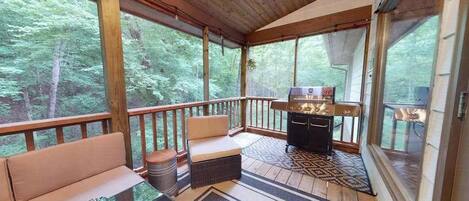 Une terrasse extérieure couverte, équipée d’un barbecue, de sièges et offrant une vue sur la forêt.