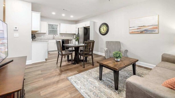 Una cocina moderna con armarios blancos, un comedor con mesa y sillas de madera, una sala de estar con sofá y mesa de centro, y un ventilador de techo.