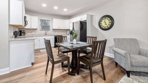 Una cocina moderna con armarios blancos, una mesa de comedor con sillas, un reloj en la pared y un sillón gris.