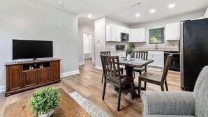 Una cocina moderna con armarios blancos, un comedor con sillas de madera, un televisor de pantalla plana y una planta en maceta.