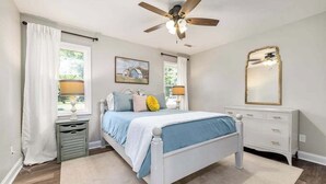 Un dormitorio con cama, ventilador de techo, cómoda, ventana con cortinas y un cuadro en la pared.