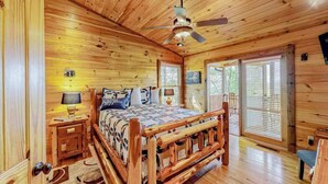 Ein Schlafzimmer einer Holzhütte mit einem Bett, Nachttischen, einem Deckenventilator und großen Fenstern.