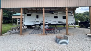 Ein überdachter Wohnmobilstellplatz mit einem weißen Wohnmobil, Kiesböden und einem Metallfass.
