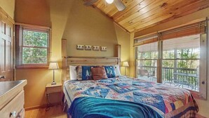 Une chambre avec un plafond en bois, un lit recouvert d’une courtepointe, une table de chevet, une commode et une fenêtre avec des stores.