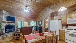 Ein Interieur einer Holzhütte mit Kamin aus Stein, einem Esstisch mit roten Platzsets, einer Küche mit Kühlschrank und Herd sowie einem an der Wand befestigten Fernseher.