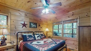 Ein Schlafzimmer einer Holzhütte mit einem Bett, einem Deckenventilator, einer Sterndeko und einem Fenster mit Blick auf Bäume.