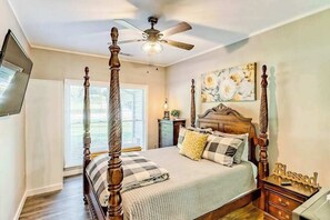 A four-poster bed with a ceiling fan, a bedside table with a 'Blessed' sign, and a floral artwork on the wall.