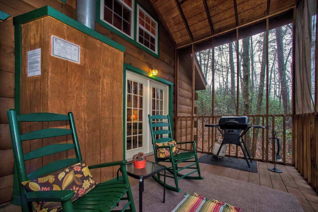 A wooden cabin with a porch, green chairs, a table, a grill, and a view of the forest.