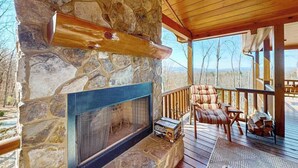A stone fireplace with a metal screen, a wooden deck with a cushioned chair, and a view of the landscape. - Bougie Bear Cabin Stay (Morganton)