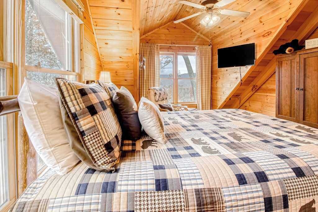 A wooden cabin interior with a bed, a ceiling fan, a television, and a window.